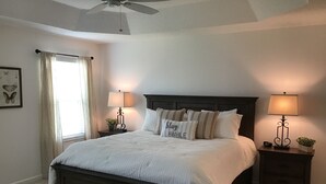 Un lit bien fait, avec une tête de lit sombre, deux tables de chevet avec des lampes, un ventilateur de plafond et une fenêtre avec des rideaux.
