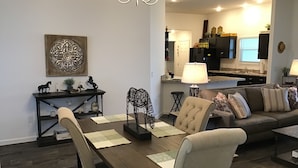 Un coin repas avec une table en bois, des chaises et un lustre. On aperçoit un canapé et un élément décoratif mural à l’arrière-plan.