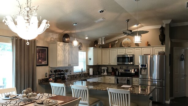 A modern kitchen with white cabinetry, stainless steel appliances, and a dining area with a wooden table and white chairs. - 1023 Blue Heron Ave (The Villages)
