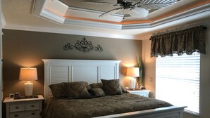 A bedroom with a white bed, two bedside tables with lamps, a ceiling fan, and a window with curtains. - 1023 Blue Heron Ave (The Villages)