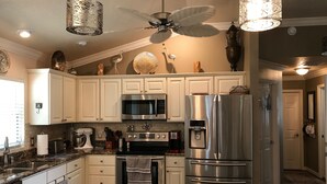 A modern kitchen with white cabinets, stainless steel appliances, granite countertops, and decorative elements like a ceiling fan and pendant lights. - 1023 Blue Heron Ave (The Villages)