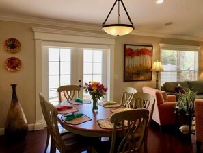 Uma sala de jantar com uma mesa redonda, cadeiras e um vaso de flores. Há um quadro na parede e um abajur em uma mesa lateral.