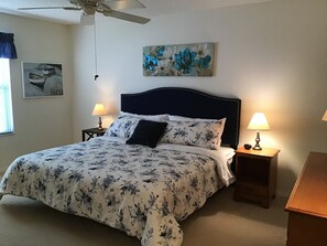 A neatly made bed with floral bedding, two bedside lamps, a ceiling fan, and a floral painting on the wall. - 1614 Gibbes Way (The Villages)