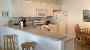 A kitchen with white cabinets, a microwave, stove, and refrigerator. There are two wooden stools and a dining table with chairs. - 1614 Gibbes Way (The Villages)