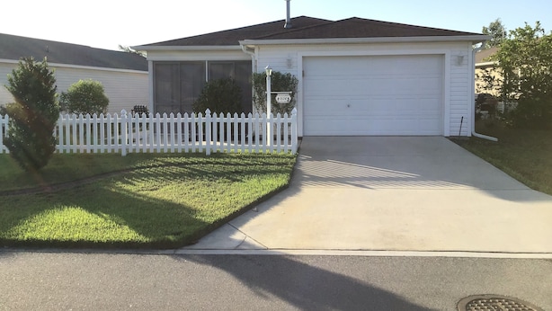 A single-story house with a white garage door, a white picket fence, and a small front yard with grass and a tree. - 1614 Gibbes Way (The Villages)