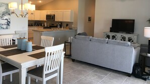 Un salon moderne avec un canapé gris, une table à manger avec des chaises blanches, un coin cuisine avec des meubles blancs, un ventilateur de plafond et un téléviseur à écran plat.