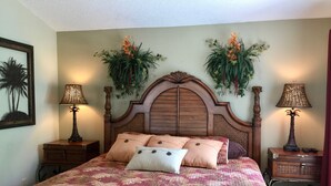 Een bed met een bloemenprint dekbed, twee nachtkastjes met lampen, en decoratieve planten aan de muur.