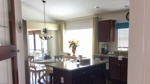 Cocina con isla central, armarios de madera, comedor con sillas, candelabro y ventana con cortinas.