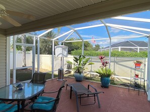 A covered patio with a glass enclosure, featuring a table and chairs, potted plants, and a metal shelf with a flag. - 2351 Bayberry Ct (The Villages)
