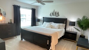 Een slaapkamer met een bed, een plafondventilator, een nachtkastje, een dressoir, een raam met gordijnen en een kamerplant.