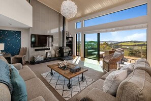 A spacious living room with a large sofa, a coffee table, and a fireplace. There is a television mounted on the wall and a large window offering a view of the outdoors.