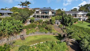 A large, modern two-story house with a spacious front yard, surrounded by lush greenery and palm trees.