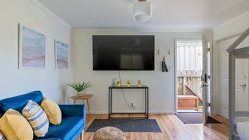 A living room with a blue sofa, a small side table with a plant, a mounted TV, and a staircase leading to an outdoor area.