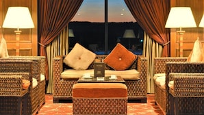 Quarto de hotel com sofá, duas luminárias, uma mesa com uma caixa e vista através de janelas amplas.