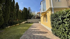 Un camino pavimentado que conduce a un edificio amarillo con un muro de piedra y altos árboles a un lado.