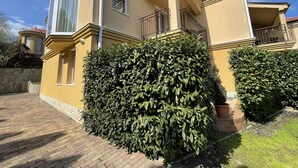 Un edificio residencial de dos pisos con fachada amarilla, un balcón con barandilla de metal y un seto bien cuidado al frente.