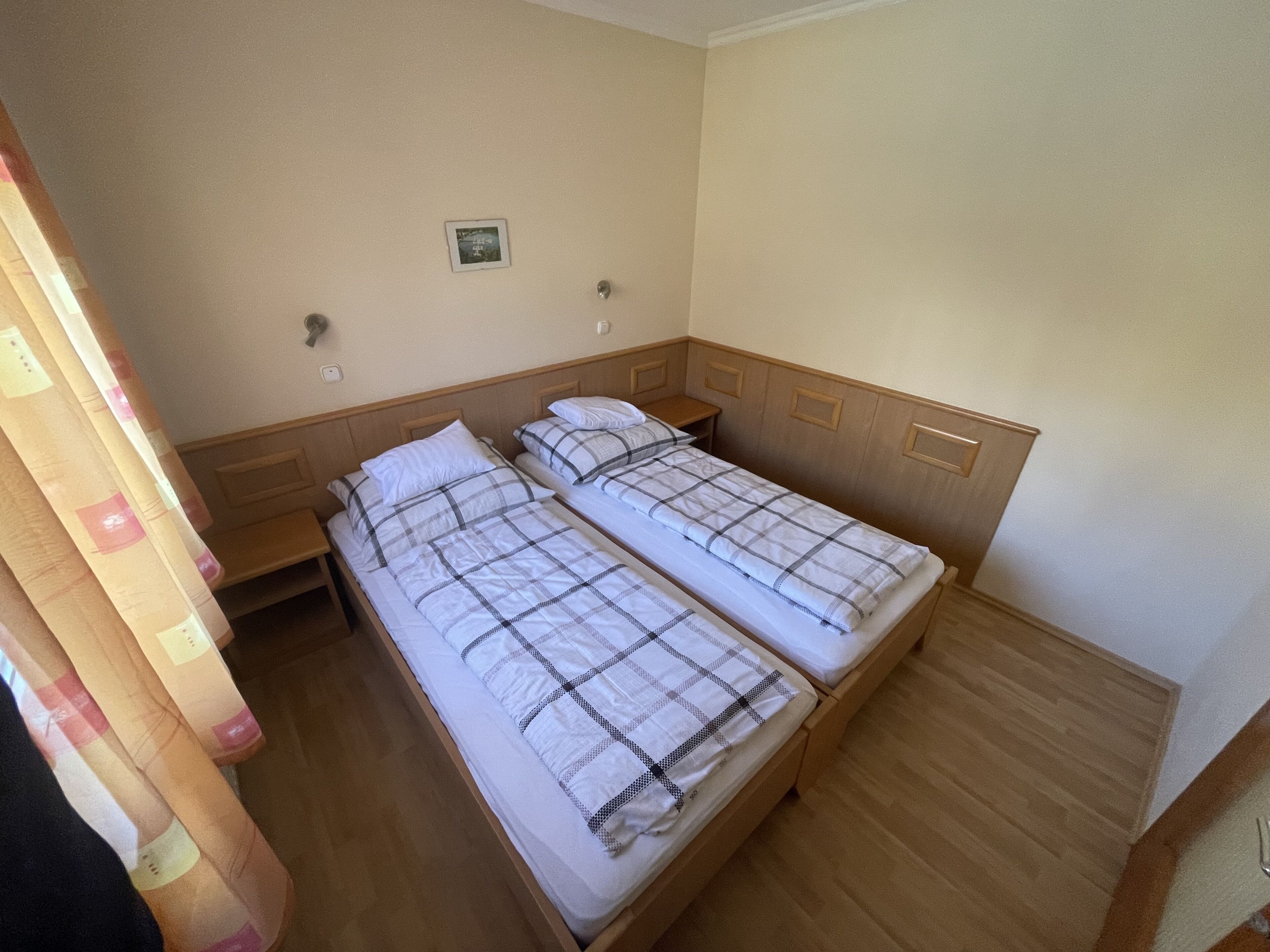 A room with two single beds, wooden headboards, a small bedside table, and a wall-mounted light switch.