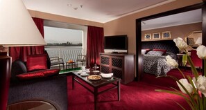Quarto de hotel com uma janela grande, cama com colcha estampada, televisão, escrivaninha com abajur e mesa de jantar com um prato de comida.