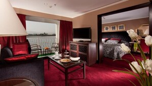 Een hotelkamer met een groot raam, een bed met een beddegoed met een patroon, een televisie, een bureau met een lamp, en een eettafel met een bord eten.