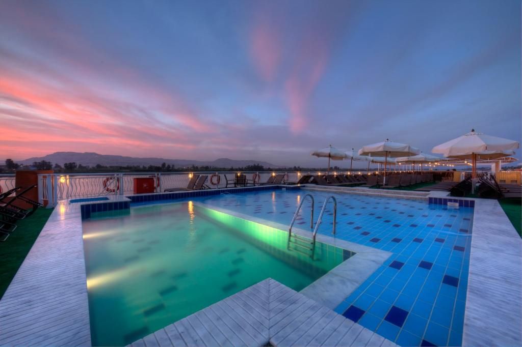 A swimming pool with clear blue tiles, surrounded by lounge chairs and umbrellas, set against a backdrop of a sunset sky.