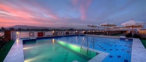 A swimming pool with clear blue tiles, surrounded by lounge chairs and umbrellas, set against a backdrop of a sunset sky.