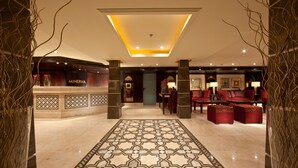 Een lobby met een vloer met geometrisch patroon, een receptiebalie met de tekst 'MINERVA' en rode zithoeken.