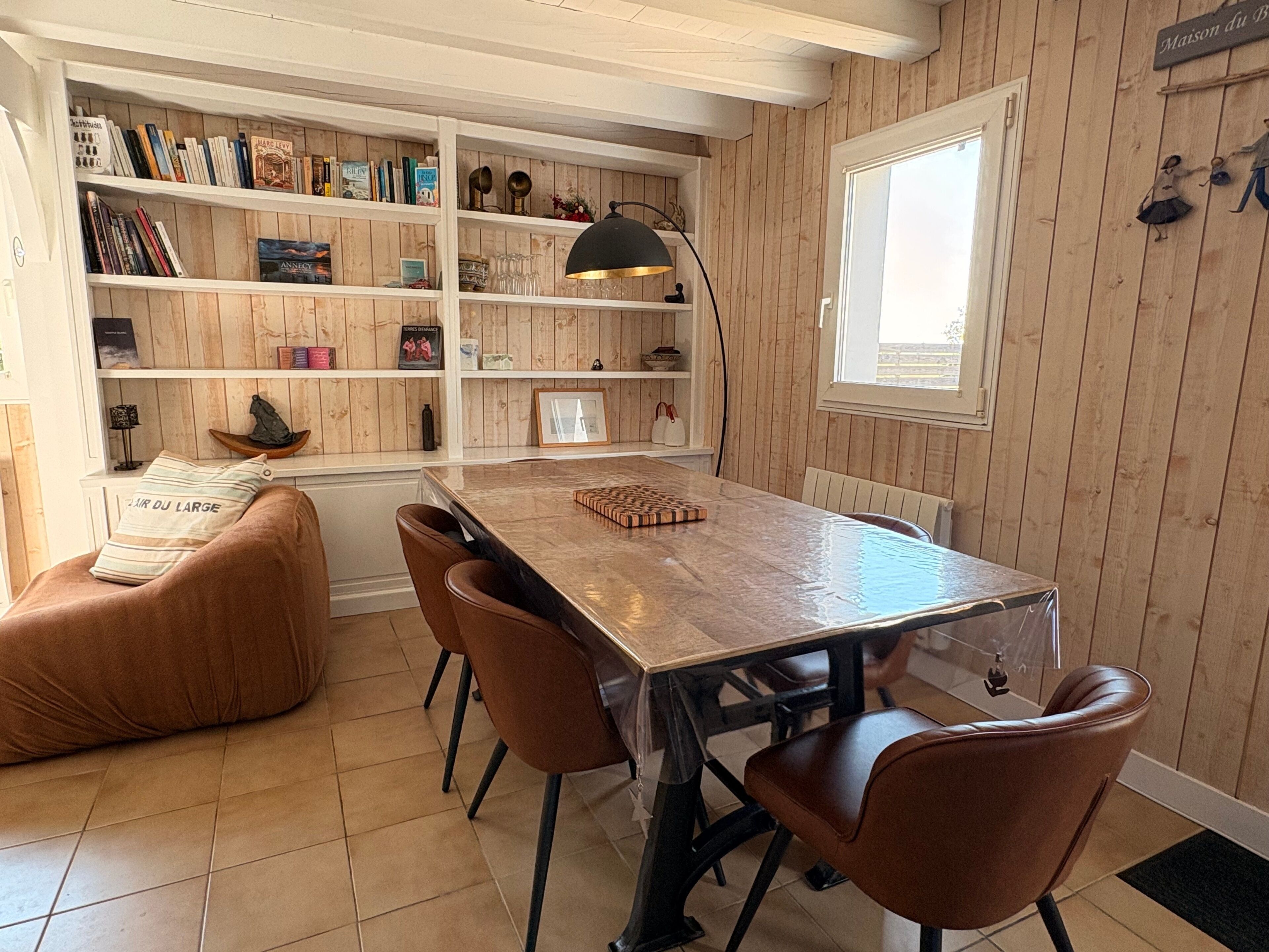 Een eethoek met een houten tafel, bruine stoelen, een boekenkast en een raam.