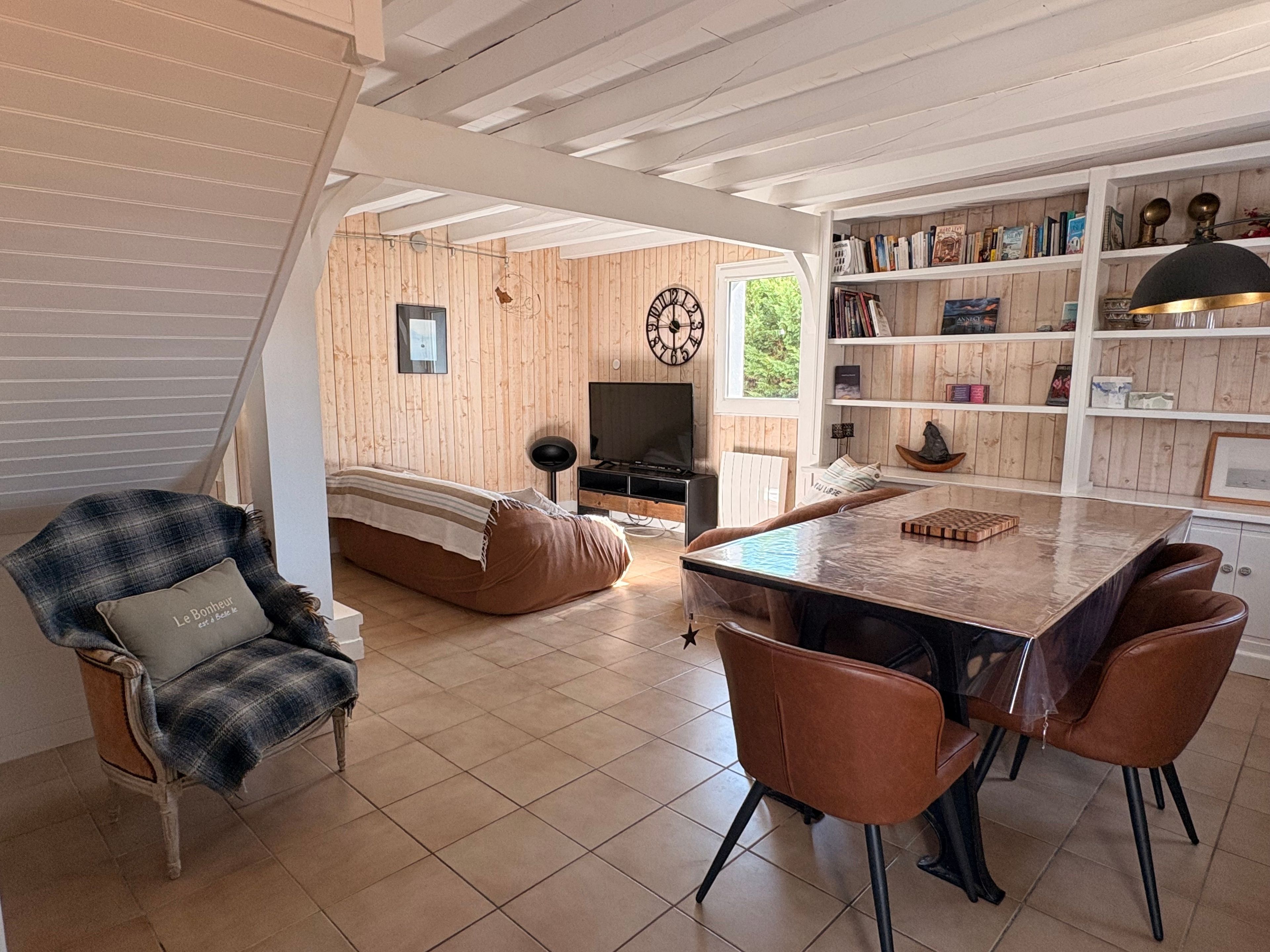 A cozy living room with a sofa, a dining table with chairs, a television, and a bookshelf filled with books and decorative items.
