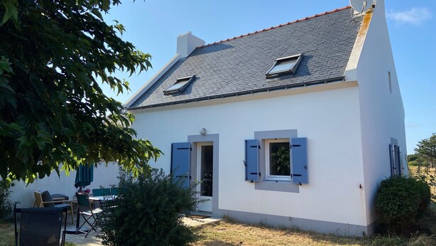 Una casa blanca con techo inclinado, dos ventanas con persianas azules y una chimenea.