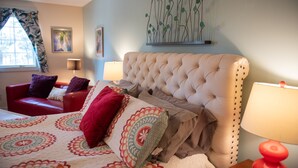 Une chambre à coucher avec un grand lit, un fauteuil rouge, une tête de lit beige et une œuvre d’art murale.