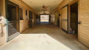 Un long couloir étroit aux murs et aux portes en bois, qui mène à une écurie où l’on aperçoit des bottes de foin et un cheval à travers une porte ouverte.