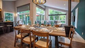 A dining area with a wooden table set for four, surrounded by chairs. There are large windows providing natural light and a view of greenery outside.