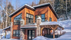 A two-story log cabin with a stone chimney, large windows, and a covered porch.