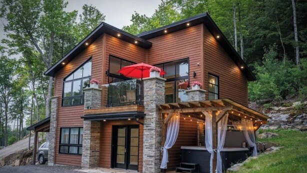 A two-story log cabin with a stone foundation, large windows, and a covered patio area with string lights.