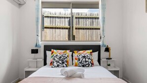 A neatly made bed with a white comforter and two patterned pillows, a bedside lamp, and a window with a view of a wooden fence. - Spacious Newly Furnished 4BD Family Retreat (Heathwood)