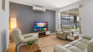 A modern living room with a flat-screen TV, two beige sofas, a wooden coffee table, and a large window with a view of a backyard. - Spacious Newly Furnished 4BD Family Retreat (Heathwood)
