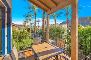 A wooden deck with a table and chairs, surrounded by a metal railing, overlooking a landscaped area with palm trees and a mountain in the background. - Desert Hideaway A | 1BR Legacy Villa (La Quinta)