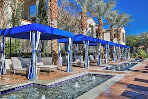 A pool area with multiple lounge chairs and umbrellas, surrounded by palm trees and a building with a tiled roof.