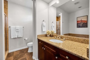 Un baño con lavamanos de encimera de granito, armarios de madera, un espejo grande y zona de ducha.