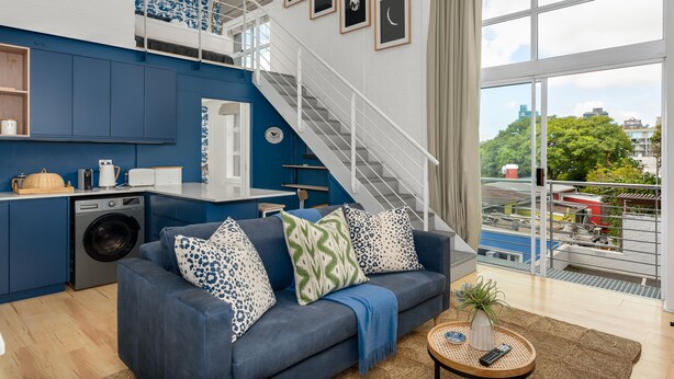 A modern living space with a blue sofa, a kitchen area, a washing machine, and a staircase leading to an upper level. - Harbour Terrace 18 (Cape Town)