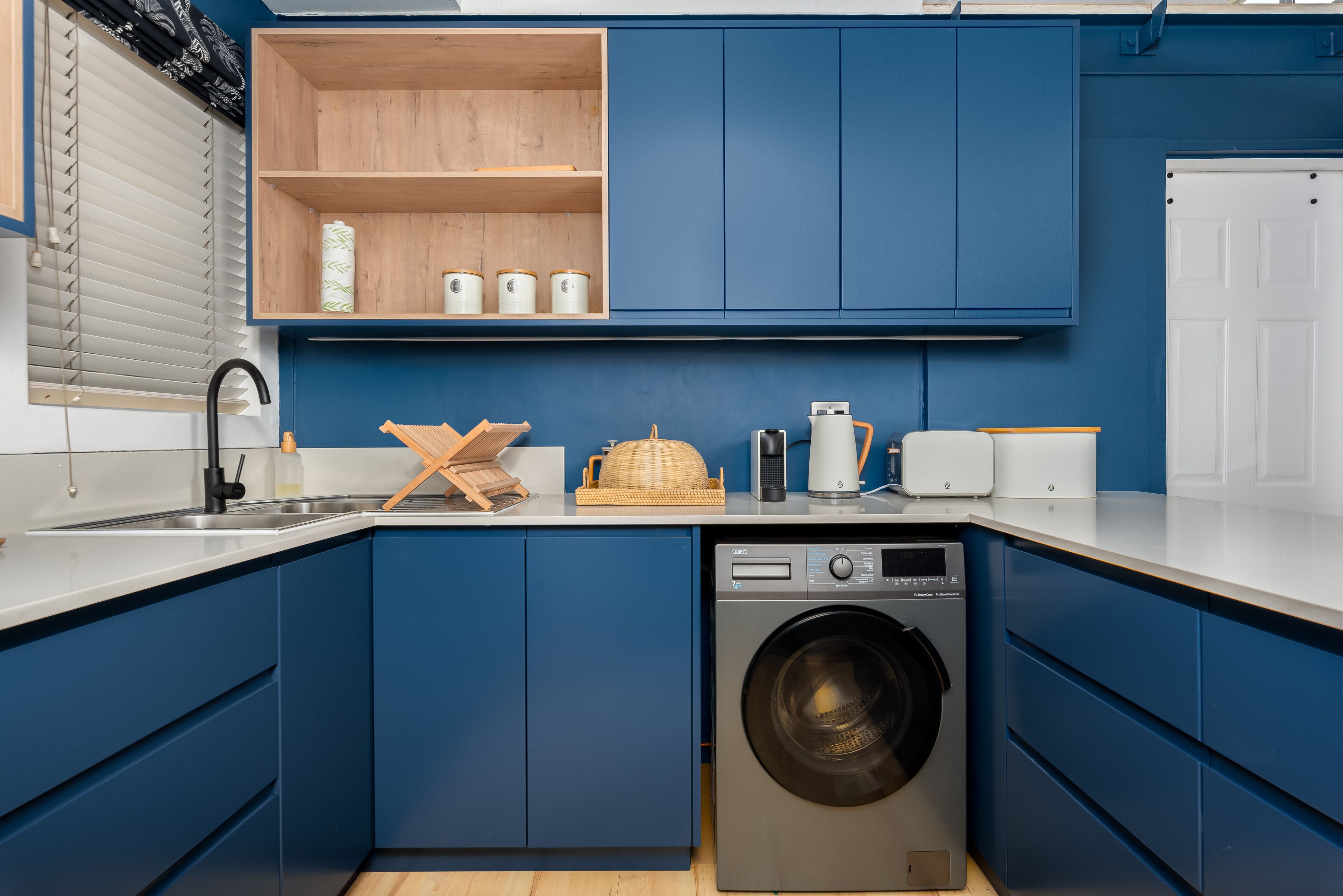 Een compacte keuken met blauwe kastjes, een wasmachine en een houten plank met keukengerei.