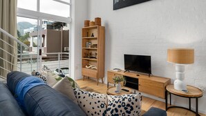 A modern living room with a large window, a wooden bookshelf, a television, and a lamp. - Harbour Terrace 18 (Cape Town)