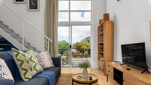 A modern living room with a staircase, a sofa with patterned cushions, a small round table with a plant, a wooden TV stand, and a large window with a view of greenery and buildings. - Harbour Terrace 18 (Cape Town)