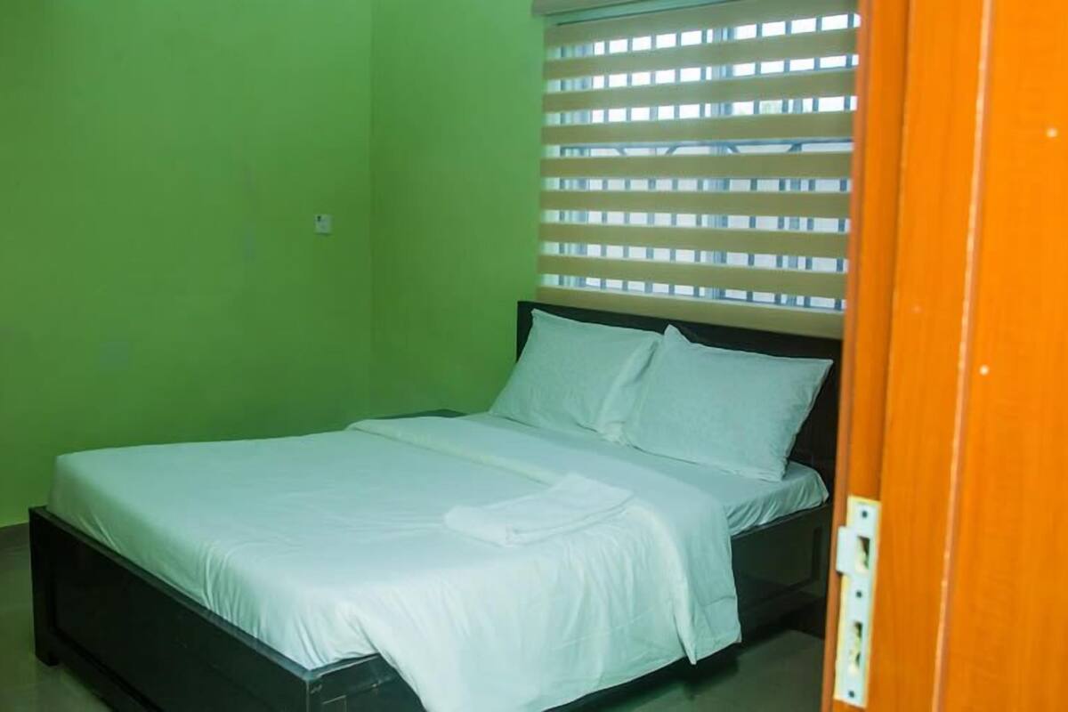 Una cama individual con ropa blanca en una habitación con paredes verdes y un ventanal con persianas.