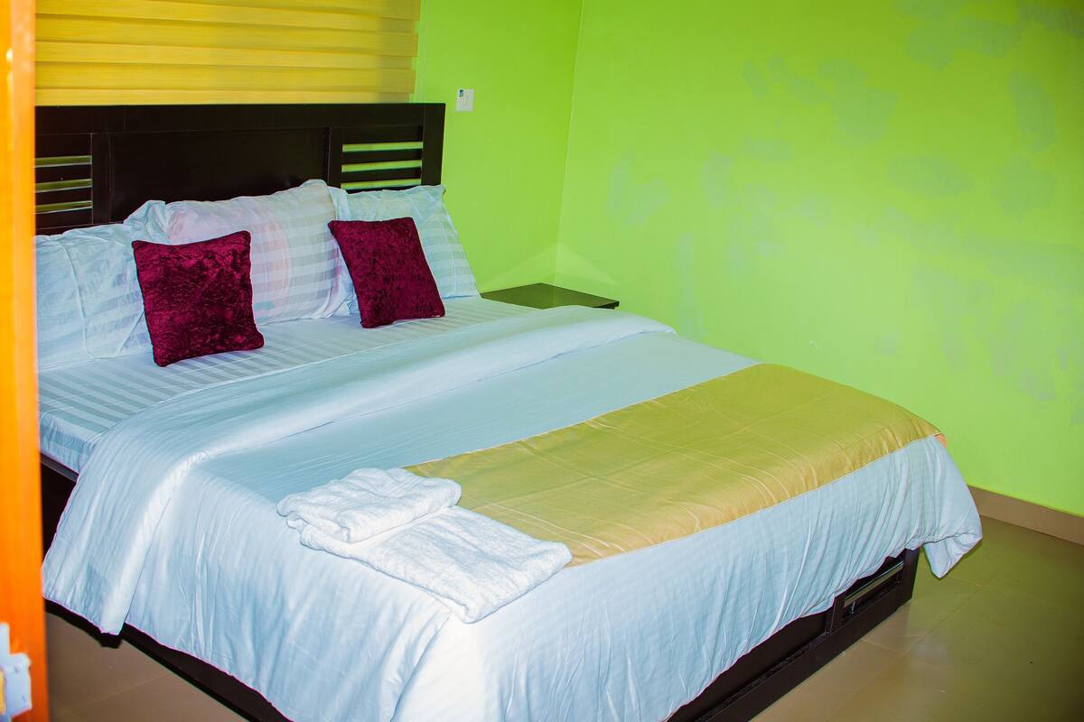 Una cama con sábanas blancas, un cojín rojo y una manta color beige.