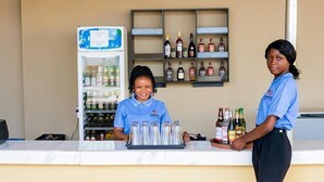 A bar area with a refrigerator stocked with beverages, a shelf with various liquor bottles, and two staff members in uniform. - Omooke Hotel (Ibadan)