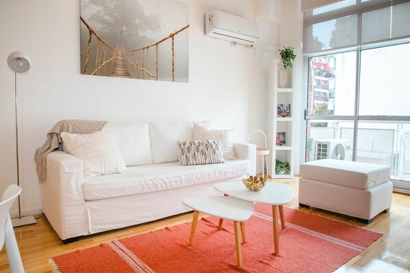 Un salon moderne avec un canapé blanc, une petite table blanche et un tapis rouge. Une grande fenêtre offre une vue sur les bâtiments à l’extérieur.