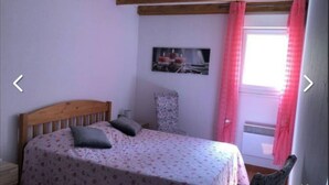 Une chambre avec un lit en bois, une literie à motifs floraux, une chaise, une petite table et une fenêtre ornée de rideaux roses.