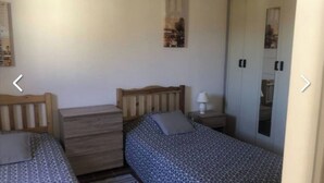 Une chambre avec deux lits simples, une commode en bois, une table de chevet avec une lampe et une armoire.