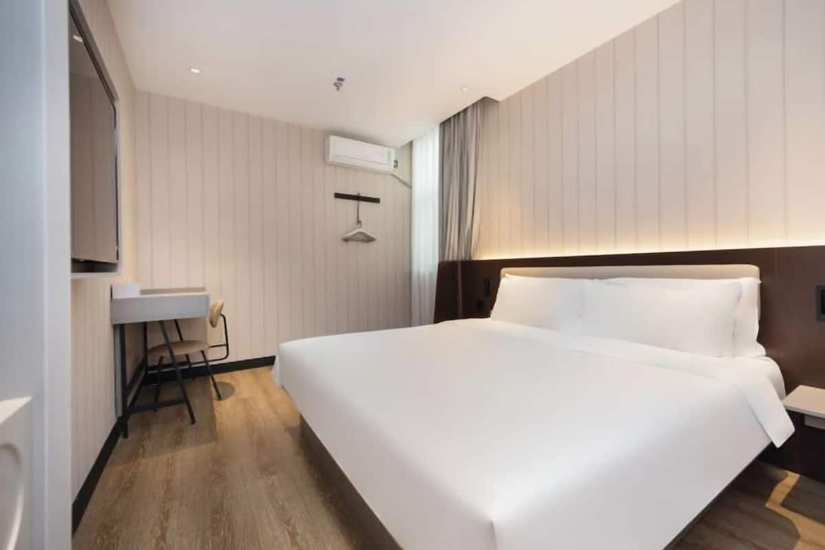 A modern hotel room with a large bed, a bedside table, a desk with a chair, and an air conditioning unit on the wall.